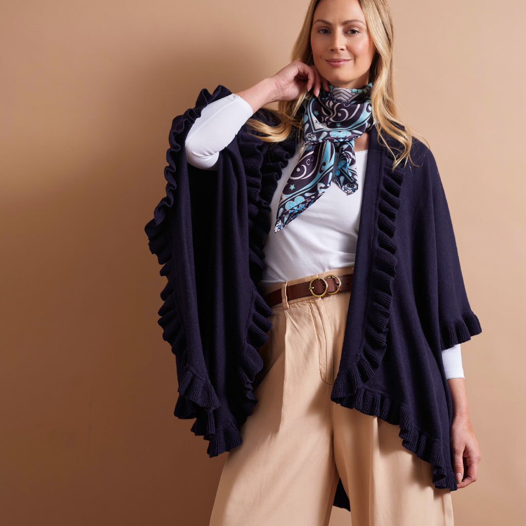 A woman models a dark navy ruffled poncho-style cardigan over a white top, beige pants, and a blue paisley neck scarf, set against a solid warm tan studio background.