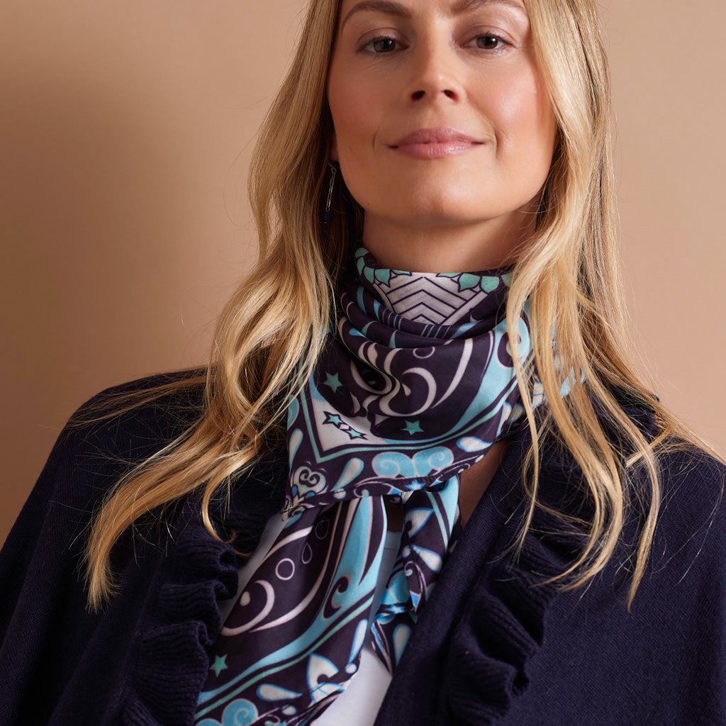 A woman with blonde hair wearing a navy blue cardigan and a patterned scarf in light blue, white, and dark navy. The background is a solid warm beige color.