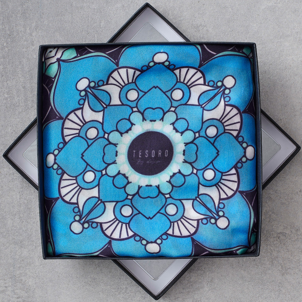 A square scarf with a vibrant blue, white, and black mandala pattern, presented inside a black gift box on a gray textured background.