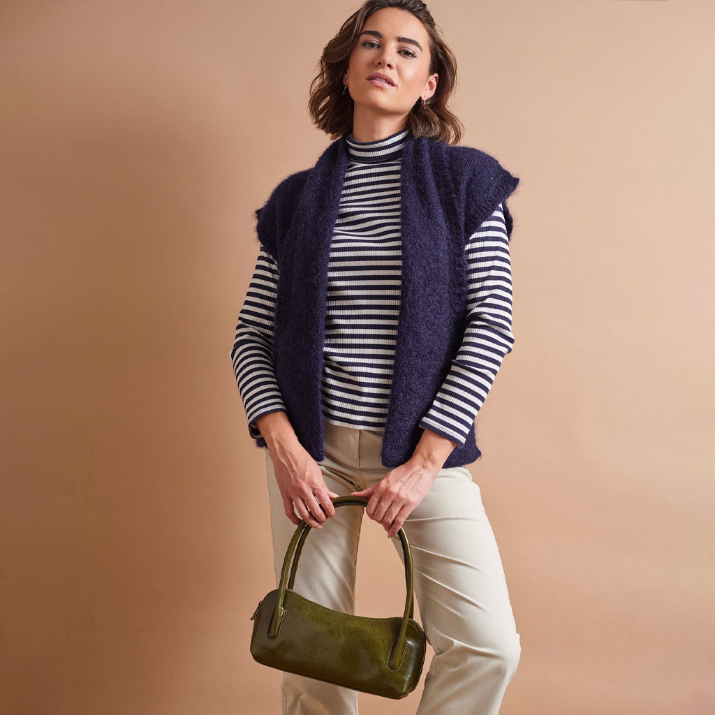 A woman models a navy blue fuzzy sleeveless vest over a navy and white striped turtleneck, paired with cream pants and holding an olive green handbag, set against a solid beige studio background.