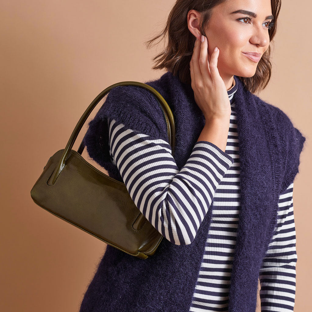 A woman wearing a navy blue fuzzy sleeveless sweater vest over a black and white striped long-sleeve top, carrying an olive green patent leather handbag over her shoulder against a solid beige background.