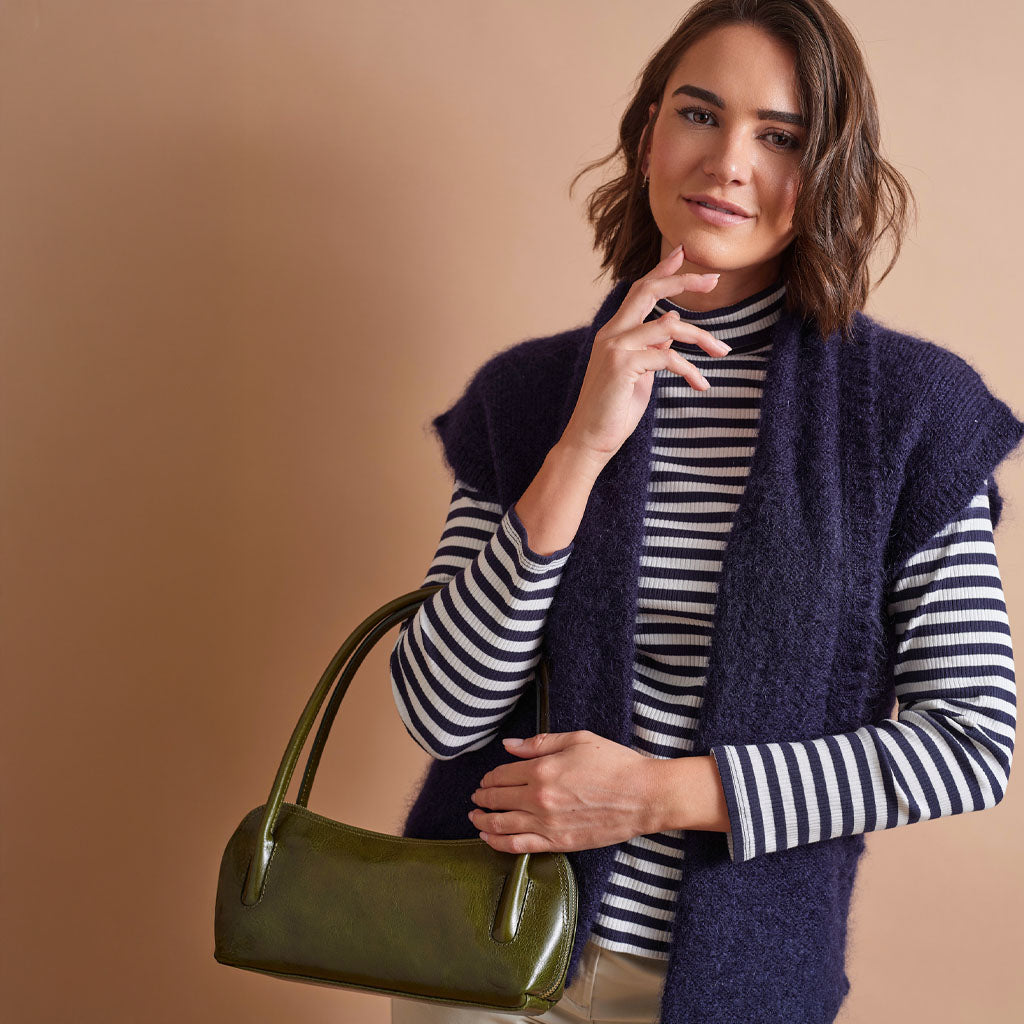 A woman models a navy blue fuzzy sleeveless vest over a navy and white striped turtleneck sweater, holding an olive green patent leather handbag against a solid warm beige background.
