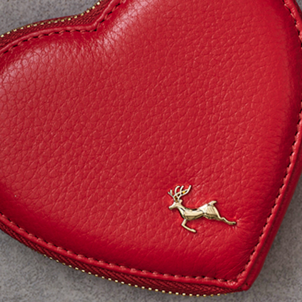 A close-up of a red textured leather heart-shaped accessory with a small gold deer emblem near the lower right and a gold zipper along the edge.