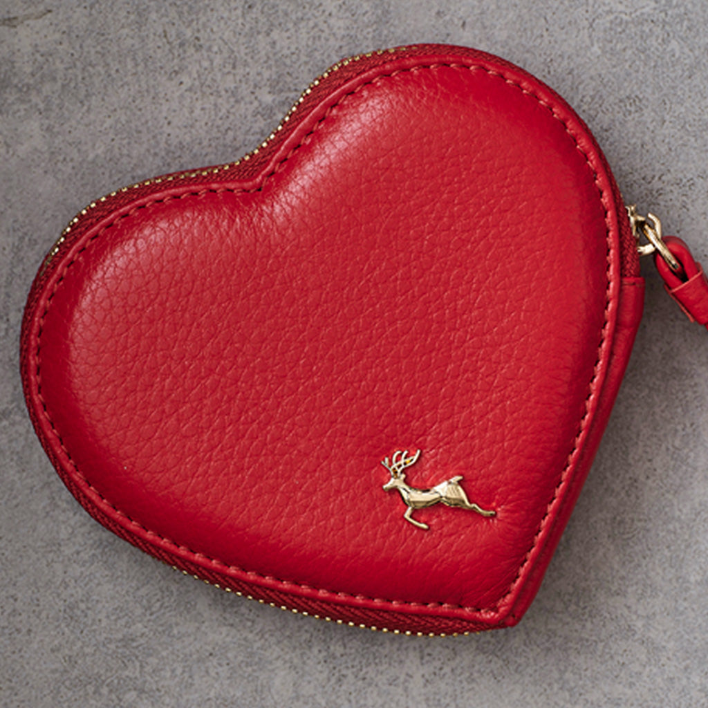 A red heart-shaped leather coin purse with a gold deer logo on the front, photographed on a textured gray background.