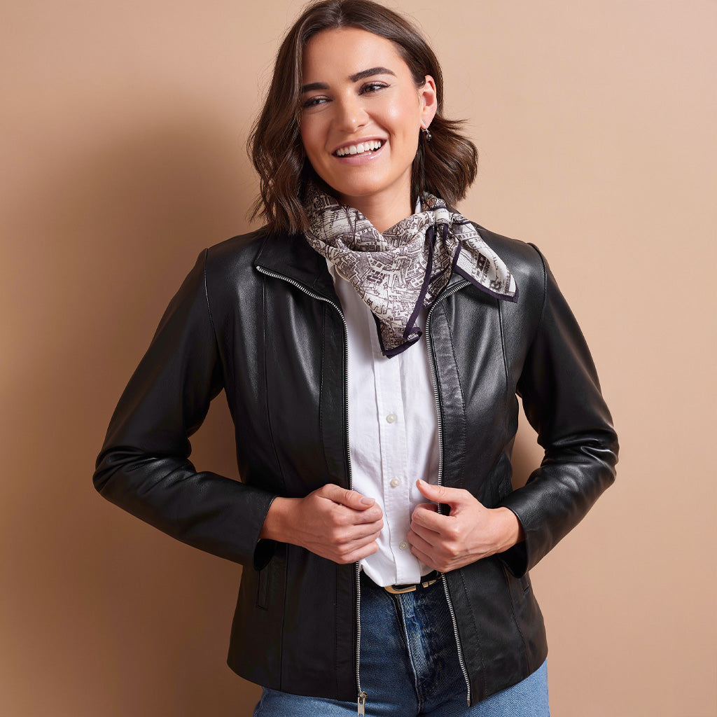 A woman wearing a black leather jacket, white button-down shirt, blue jeans, and a patterned scarf, standing against a solid tan background.
