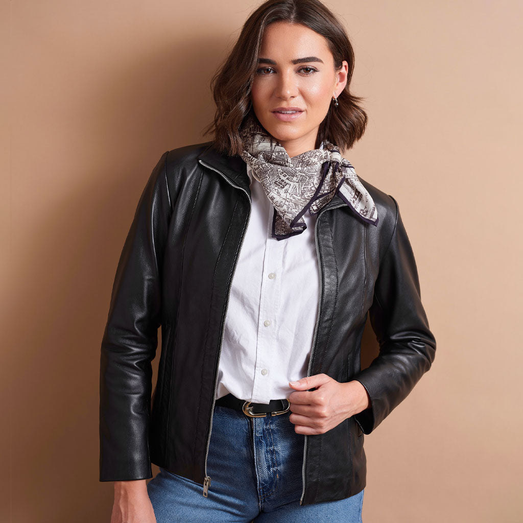 A woman wearing a black faux leather moto jacket, a white button-down shirt, blue jeans, and a patterned bandana scarf, standing against a solid beige studio background.