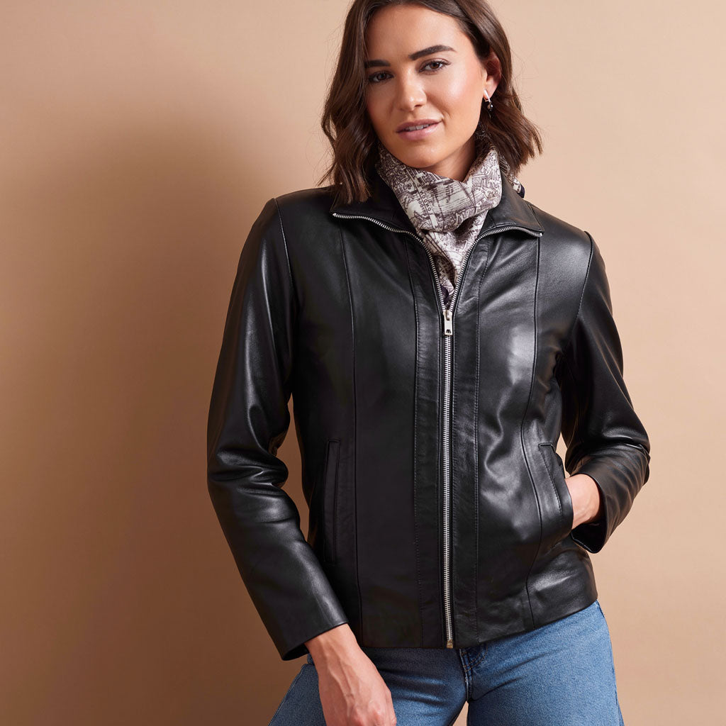 A woman wearing a black faux-leather zip-up jacket with a stand collar, paired with a gray patterned scarf and blue jeans, standing against a solid beige background.