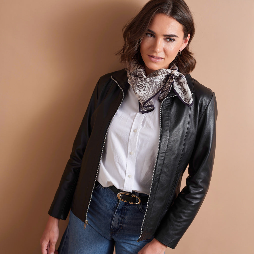 A woman wearing a black leather jacket over a white button-up shirt, styled with a patterned scarf and blue jeans, posing against a solid beige background.