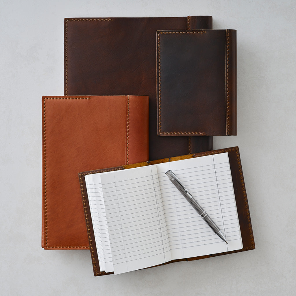 Four leather notebook covers in dark brown, medium brown, and tan are arranged on a light gray background, with one open notebook showing lined pages and a silver pen resting inside.