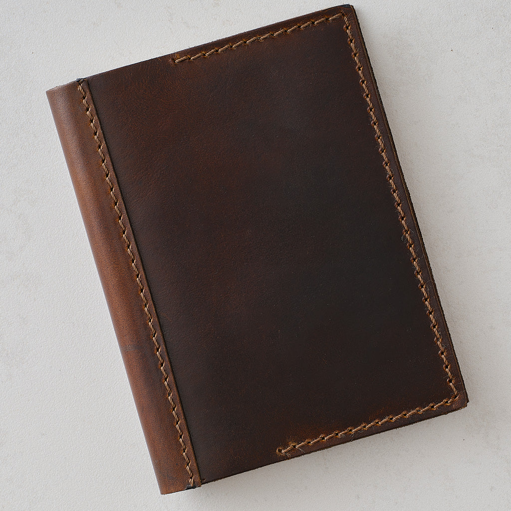 A dark brown leather notebook or journal cover with light tan contrast stitching, shown from an overhead angle on a light gray background.