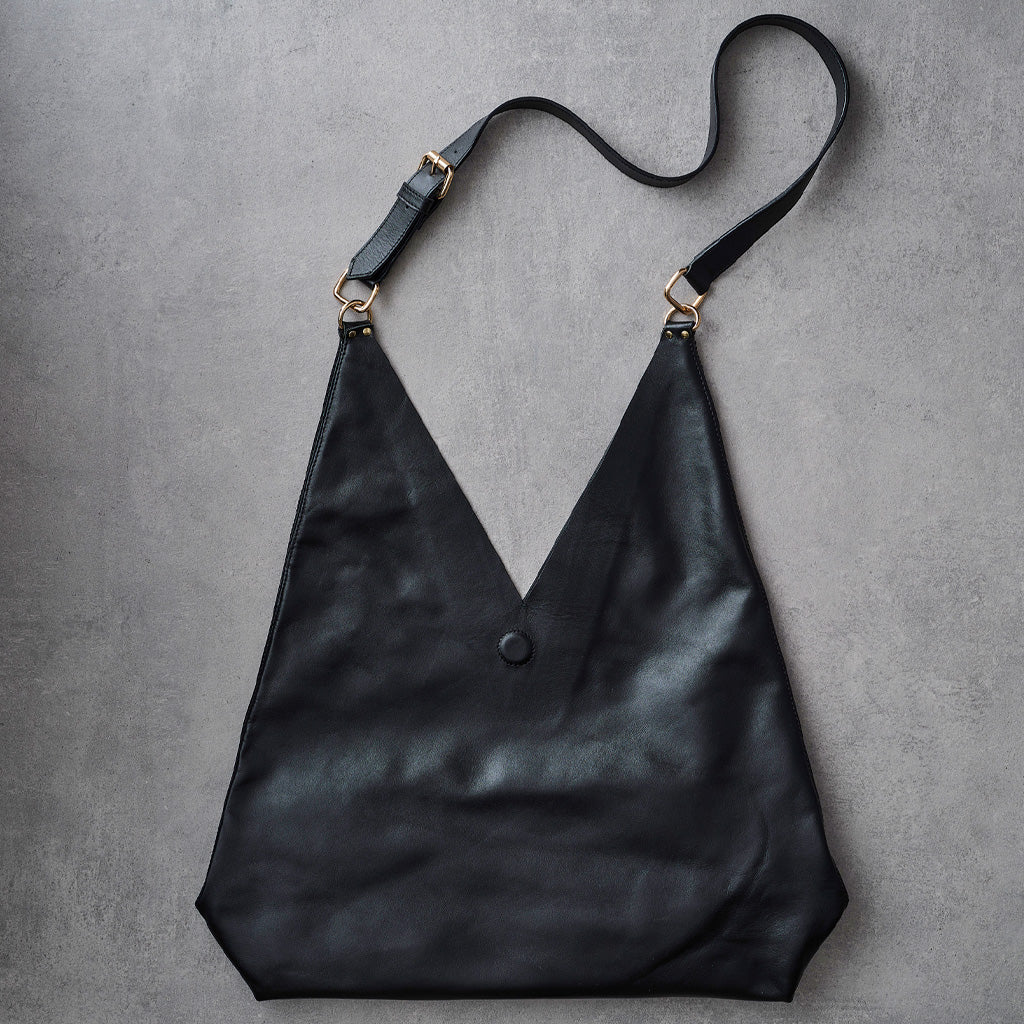 A black leather hobo-style shoulder bag with a wide body, V-shaped neckline, a single button closure, and a thin adjustable strap with gold-tone hardware, laid flat on a textured gray background.