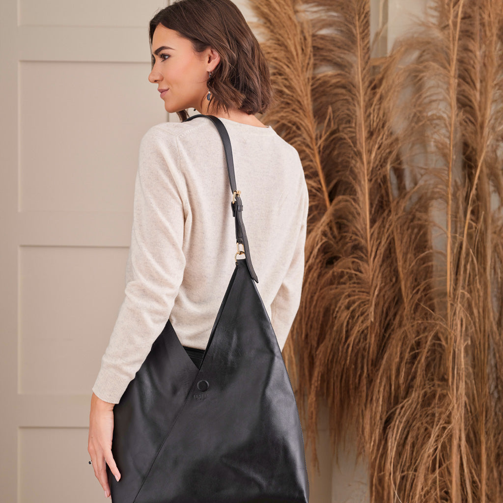 A woman wearing a cream knit sweater is shown from the back, carrying a large black leather hobo shoulder bag. The setting features a neutral beige paneled wall and a decorative arrangement of dried pampas grass on the right.