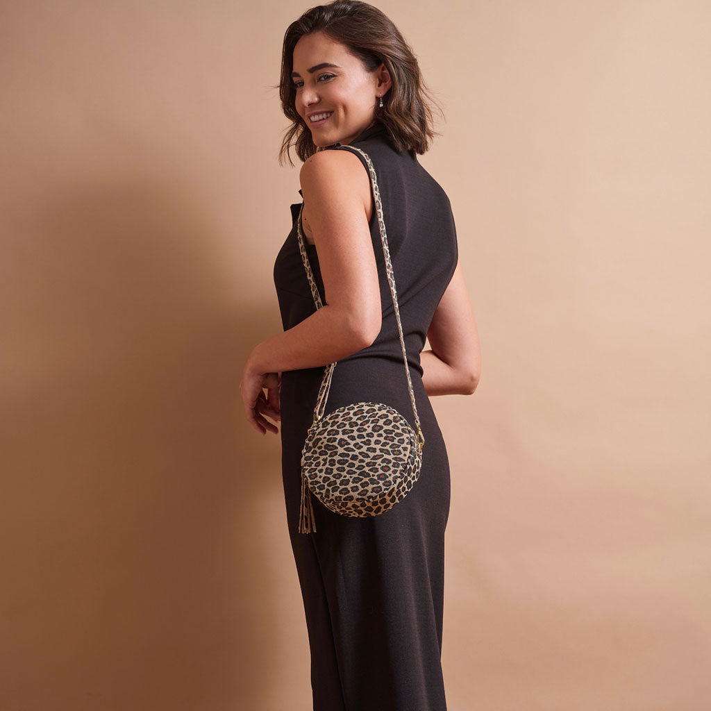 A woman in a black sleeveless dress models a round leopard-print crossbody bag with a gold-tone chain strap against a solid beige studio background.