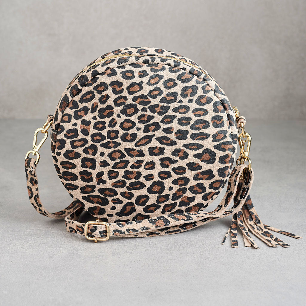 A round leopard-print crossbody bag with a matching adjustable strap and tassel detail, shown on a neutral gray background.