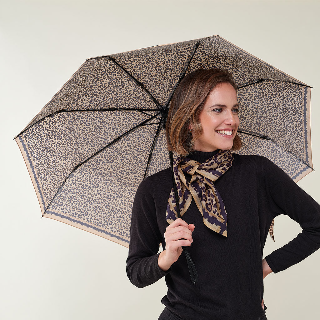 A woman holding a leopard-print umbrella and wearing a matching leopard-print scarf, dressed in a black long-sleeve top against a plain light background.