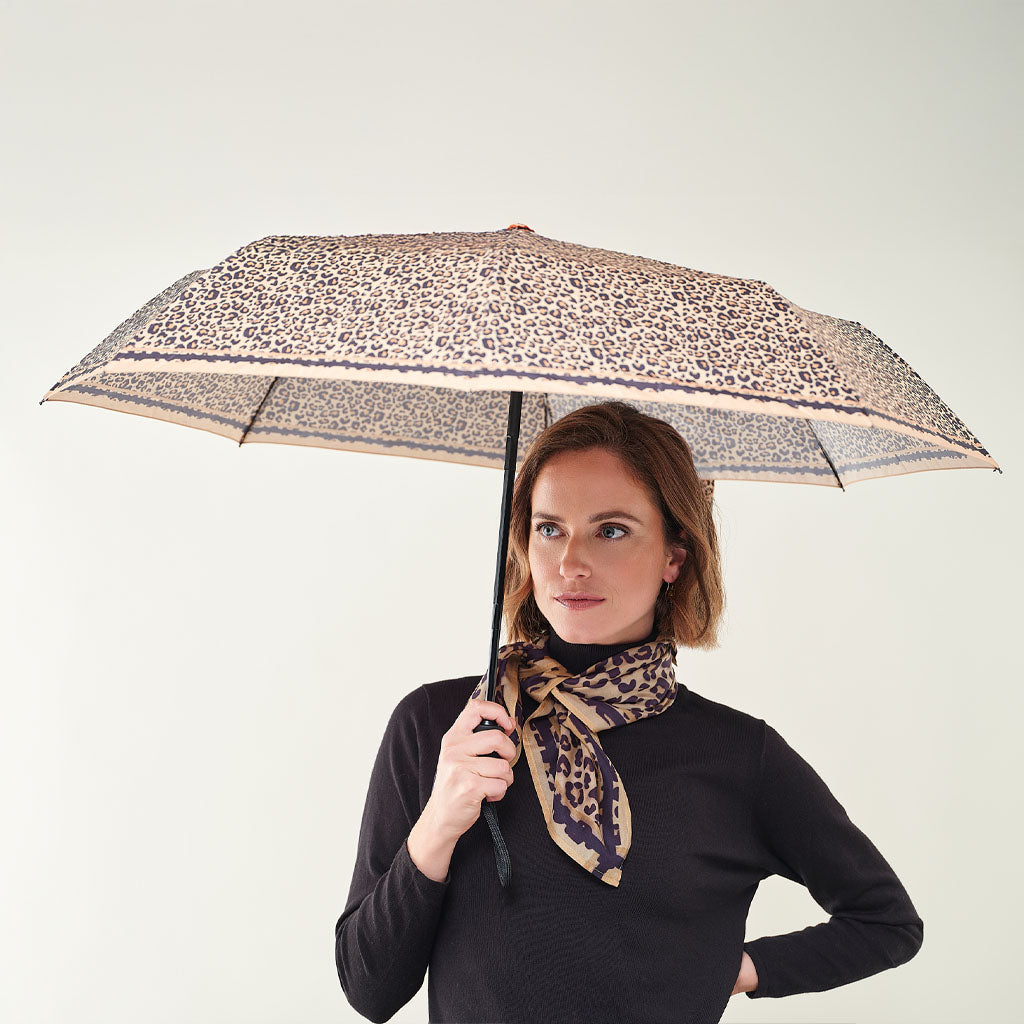 A woman holding a leopard-print umbrella with a matching leopard-print neck scarf, wearing a black long-sleeve top against a plain light gray background.