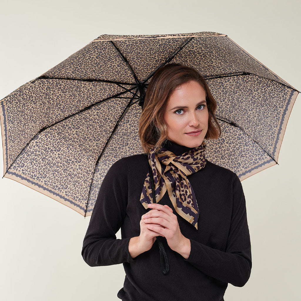 A woman holding an open leopard-print umbrella, wearing a matching leopard-print scarf and a black long-sleeve top, against a plain light beige background.