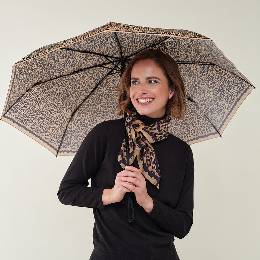 A woman holding an open leopard-print umbrella, wearing a matching leopard-print scarf and a black long-sleeve top, against a plain light beige background.