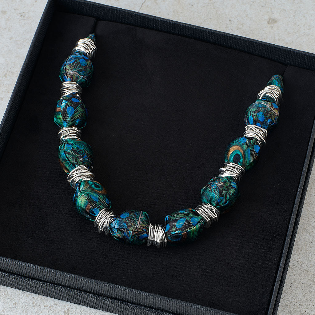 A beaded necklace with large marbled beads in teal, blue, and black, separated by silver wire-wrapped accents, displayed inside a black presentation box.
