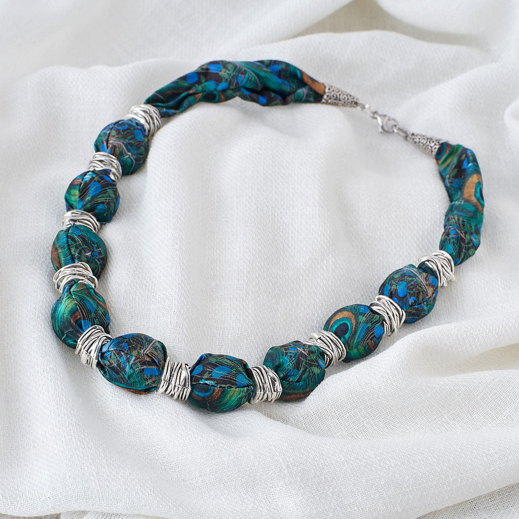 A beaded necklace with textured, irregular beads featuring a vibrant blue, green, and brown marbled pattern, separated by silver wire-wrapped spacers, displayed on a draped white fabric background.
