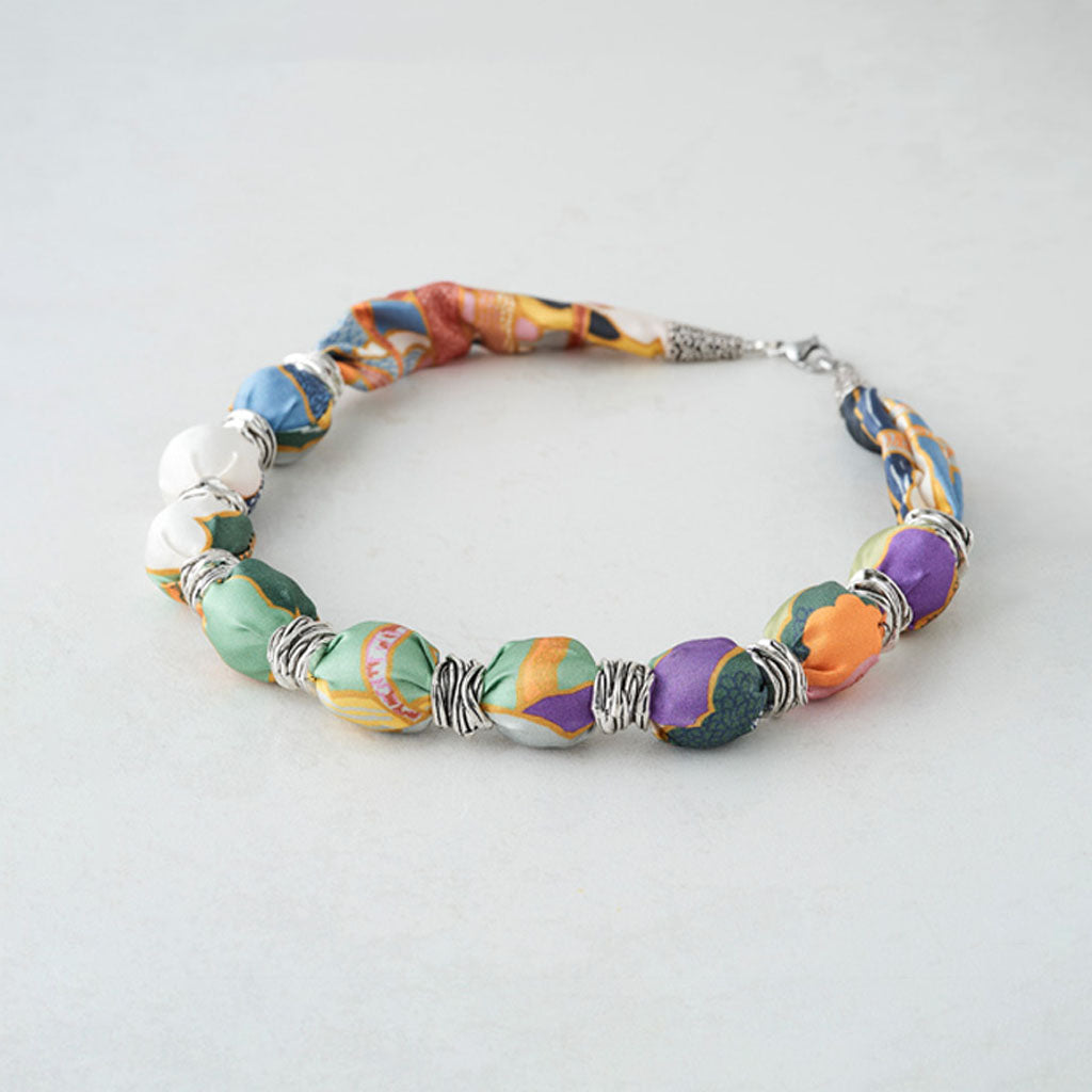 A handmade beaded bracelet featuring colorful, marbled ceramic beads in purple, green, orange, yellow, and blue, separated by silver wire-wrapped spacers and a silver clasp, shown on a light gray background.