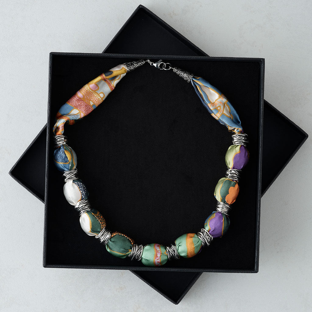 A handmade beaded necklace with colorful marbled glass beads and silver wire spacers, displayed inside an open black presentation box on a light gray surface.