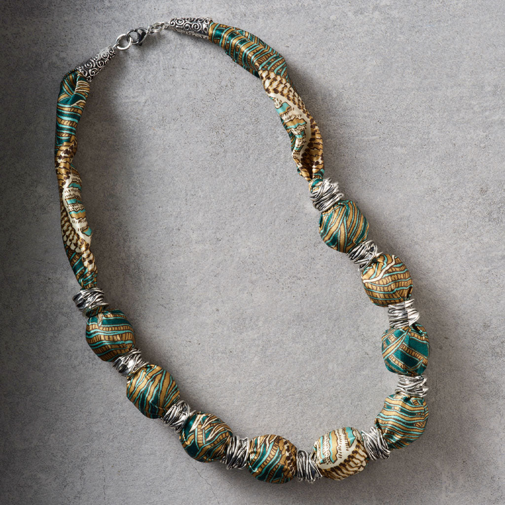 A handmade beaded necklace featuring textured, patterned paper beads in green, gold, and brown, separated by silver wire spacers, displayed on a neutral gray background.