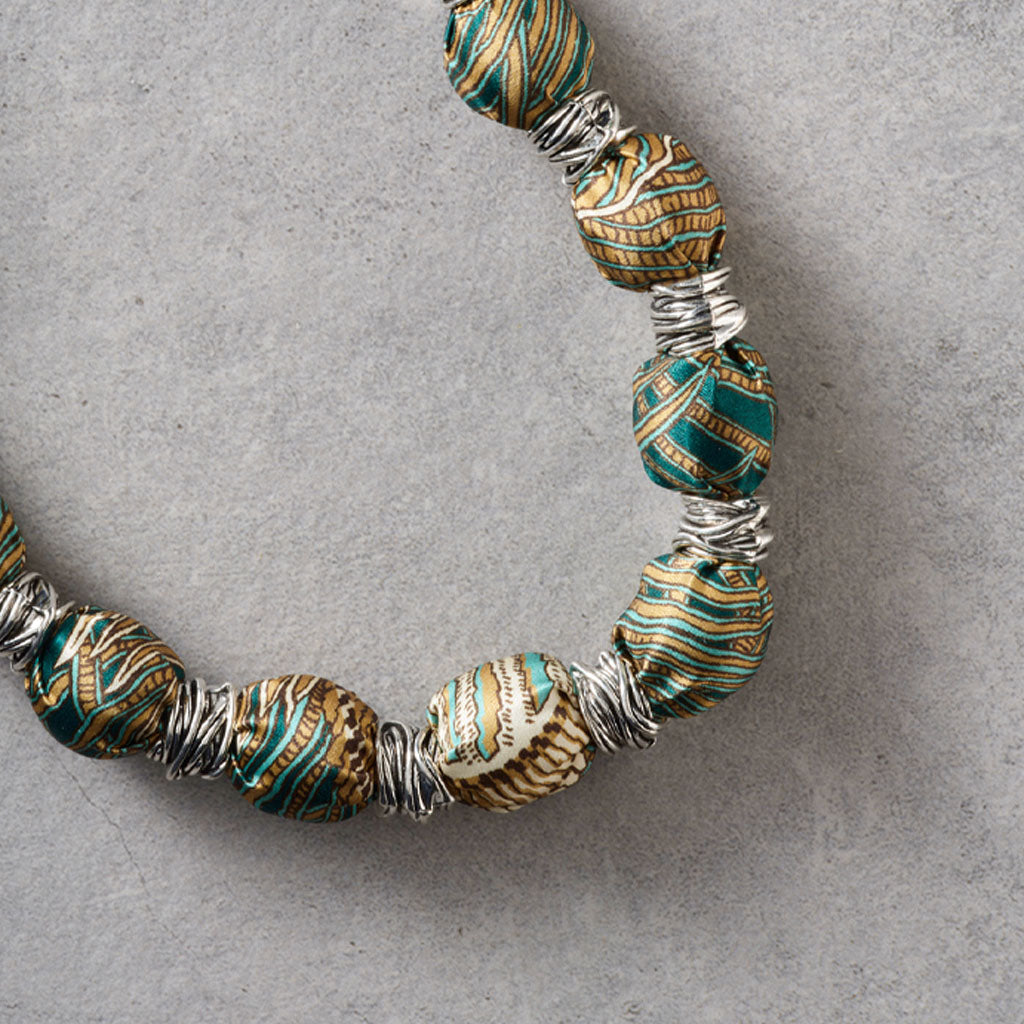 A close-up of a beaded necklace with patterned teal, gold, and white beads separated by silver wire wraps, laid on a textured gray surface.