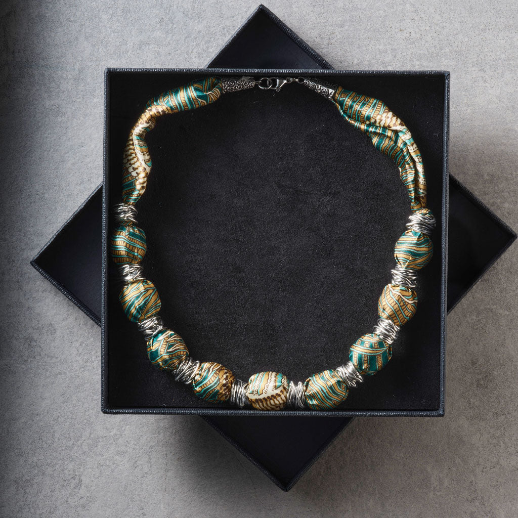 A beaded necklace with metallic gold and teal-green striped beads, displayed inside an open black jewelry box on a gray surface.