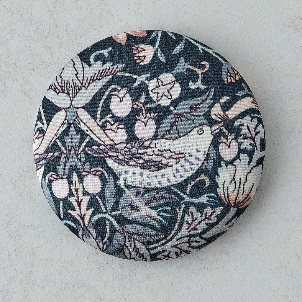 A round button pin featuring a William Morris-style floral and bird pattern in navy, gray, blush pink, and white, photographed on a light gray textured surface.