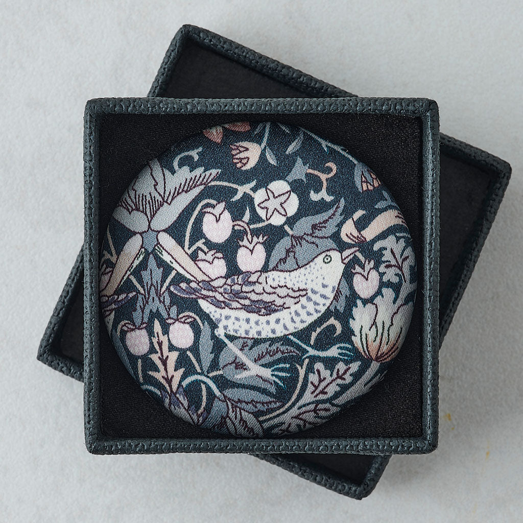 A round brooch featuring a William Morris-style bird and floral pattern in muted blue, pink, and white, presented inside an open dark textured gift box on a light gray background.