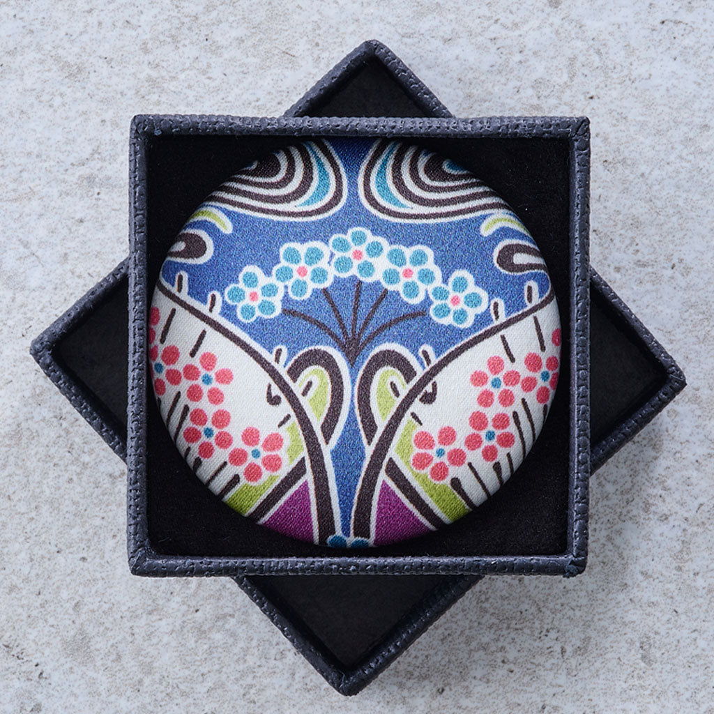 A round pinback button featuring a colorful Japanese-style floral and tree pattern, presented inside a dark gray star-shaped jewelry box on a light gray textured surface.