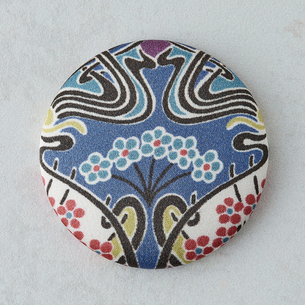 A round fabric pin or button featuring a colorful Art Deco style pattern with blue, white, black, yellow, and red details, set against a light gray textured background.