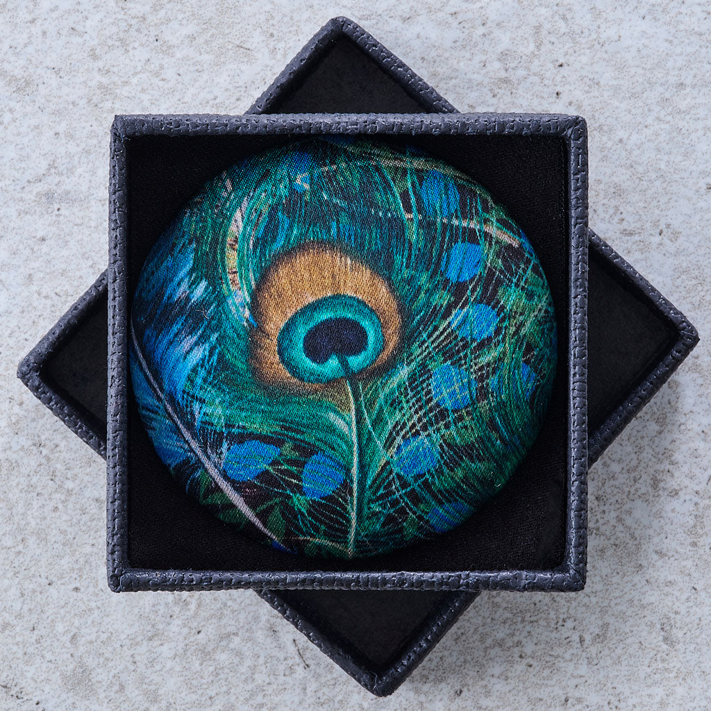A round pin featuring a detailed peacock feather design in shades of teal, blue, green, and gold, displayed inside a black star-shaped jewelry box on a light gray textured background.