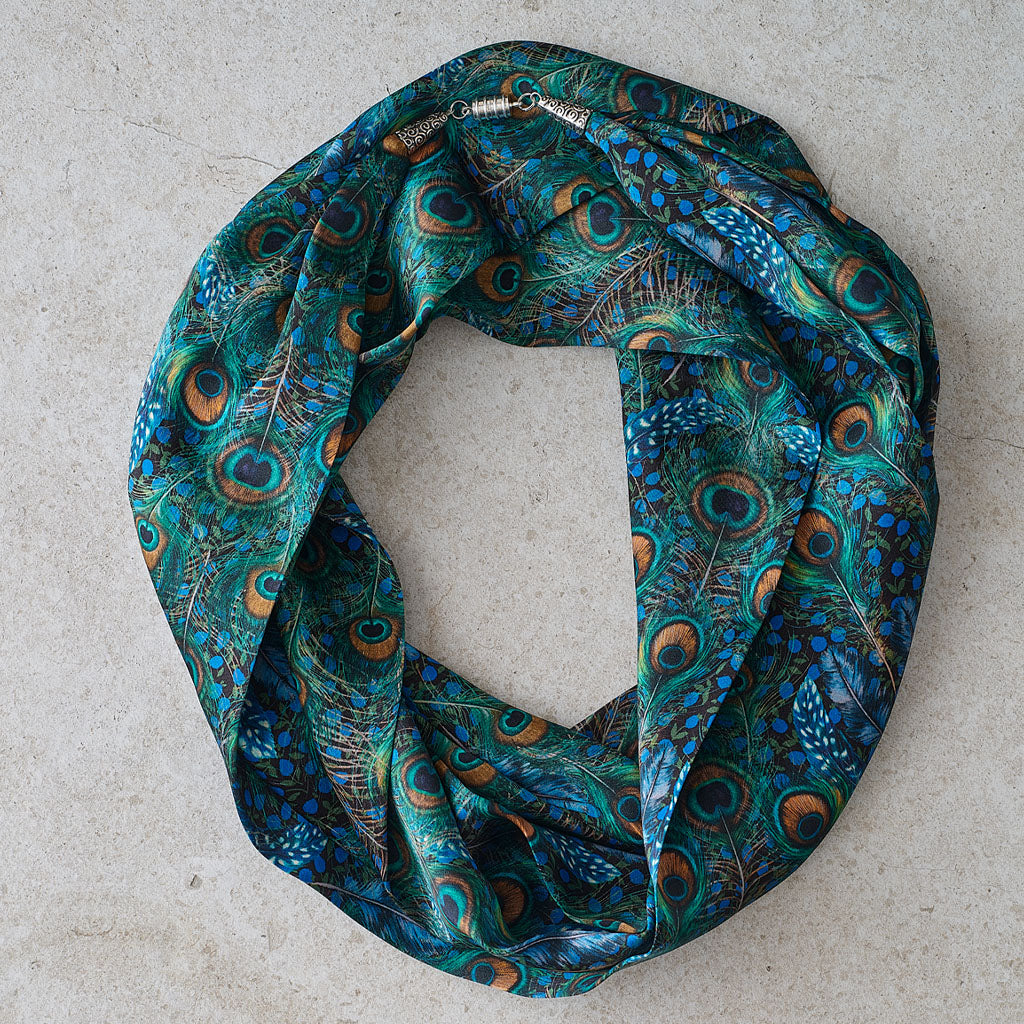 A peacock feather print infinity scarf in shades of teal, blue, and brown, displayed in a loop on a light gray textured surface.
