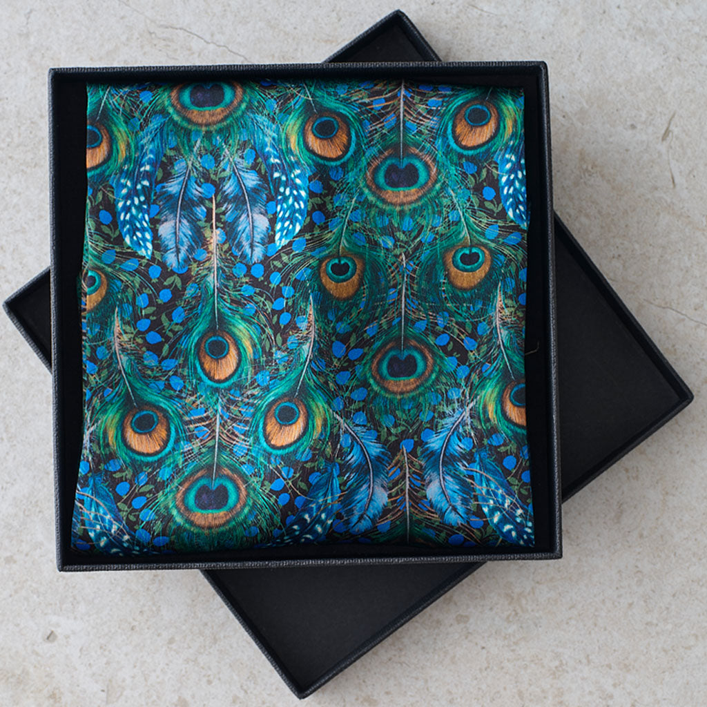 A square black gift box is open to reveal a folded fabric item with a vibrant peacock feather print in shades of teal, blue, green, and gold. The box is placed on a light beige stone surface.