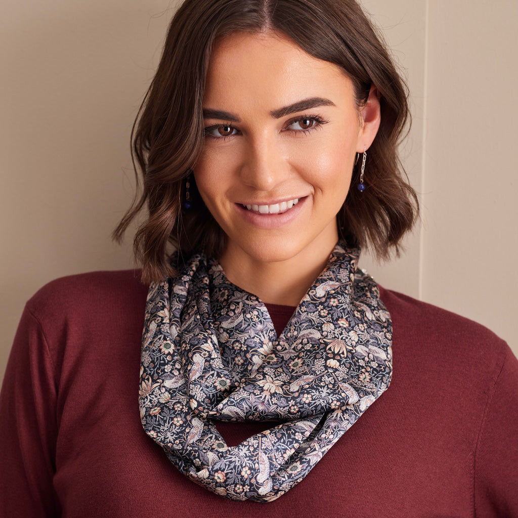 A smiling woman with shoulder-length brown hair models a patterned infinity scarf. The scarf features a detailed floral and paisley design in navy blue, beige, and white.