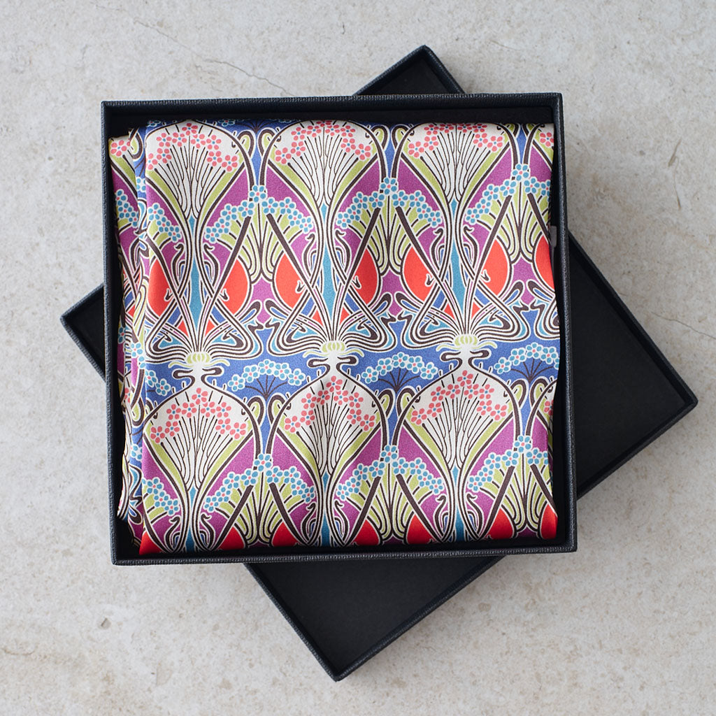 A folded square scarf with a vibrant Art Deco pattern in blue, pink, red, and gold, presented inside an open black gift box on a light stone surface.