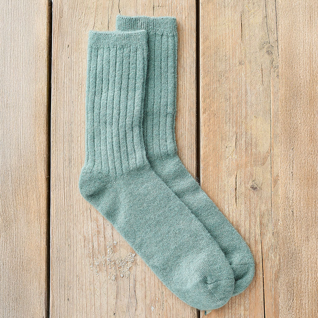 A pair of ribbed knit crew socks in a muted sage green color, laid flat on a light brown wooden plank background.