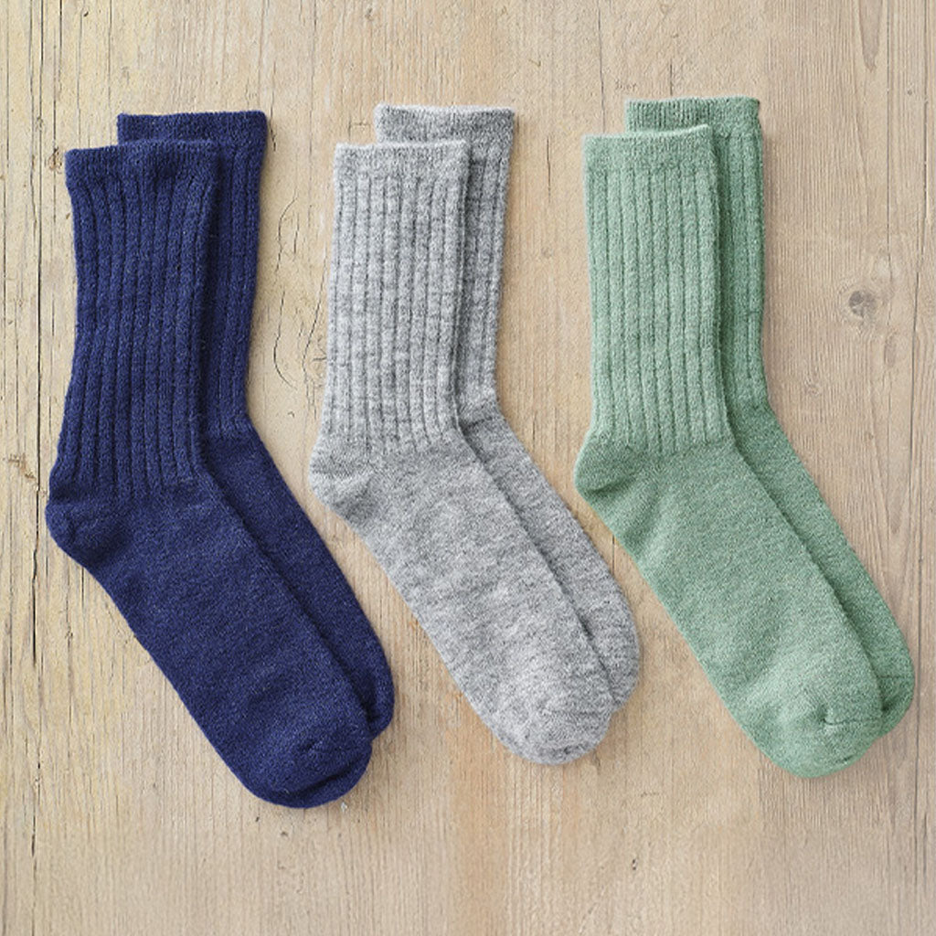 Three pairs of ribbed crew socks laid flat on a light wood surface: navy blue, heather gray, and sage green.