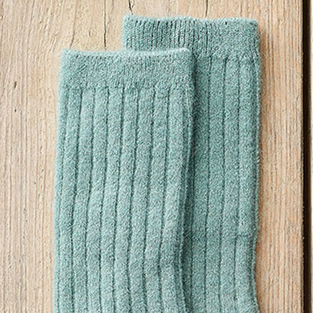A pair of ribbed knit socks in a dusty sage green color, displayed on a light wood background.