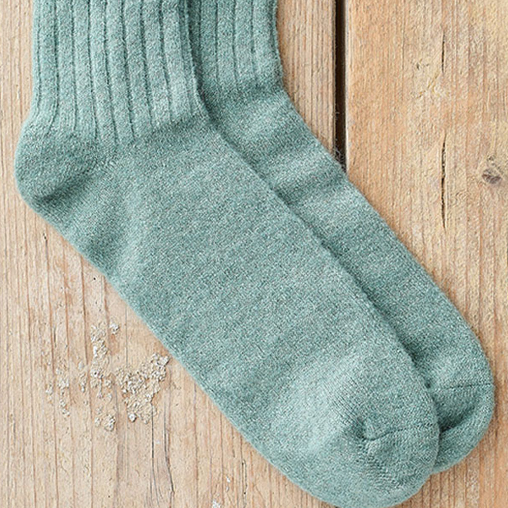 A pair of ribbed knit crew socks in a muted sage green color, laid flat on a light brown wooden surface.