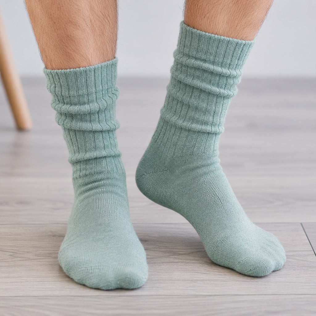 A pair of ribbed crew socks in a muted sage green color, worn on a person's feet standing on a light wood floor.