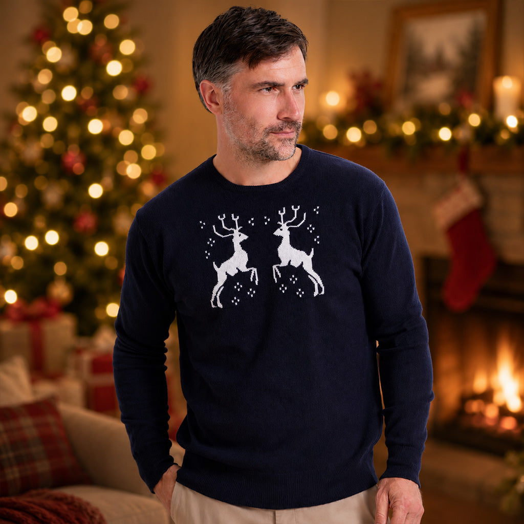 A man wearing a cashmere navy blue crewneck sweater with a white graphic of two leaping reindeer, standing in a cozy, warmly lit living room with a Christmas tree and fireplace in the background.