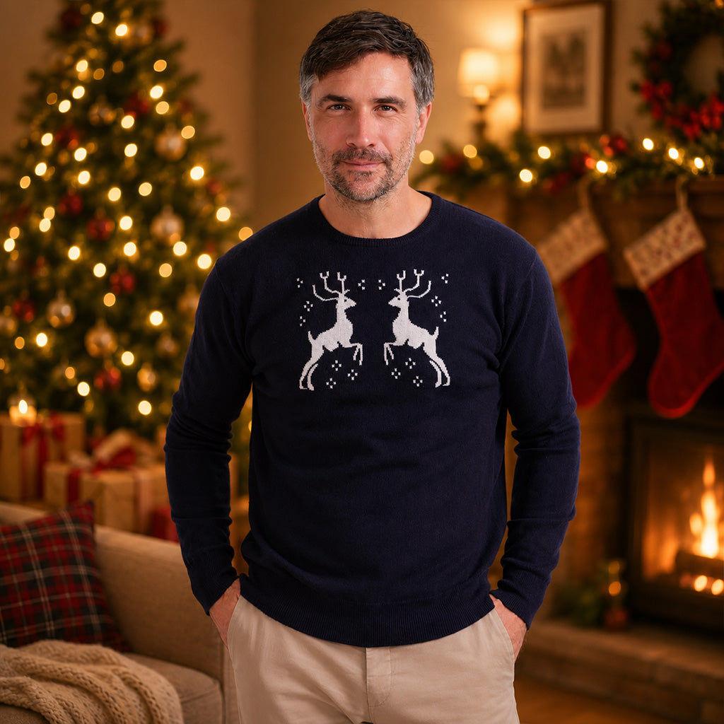 A man wearing a cashmere navy blue crewneck sweater with a white graphic of two leaping reindeer, standing in a cozy, warmly lit living room with a Christmas tree and fireplace in the background.