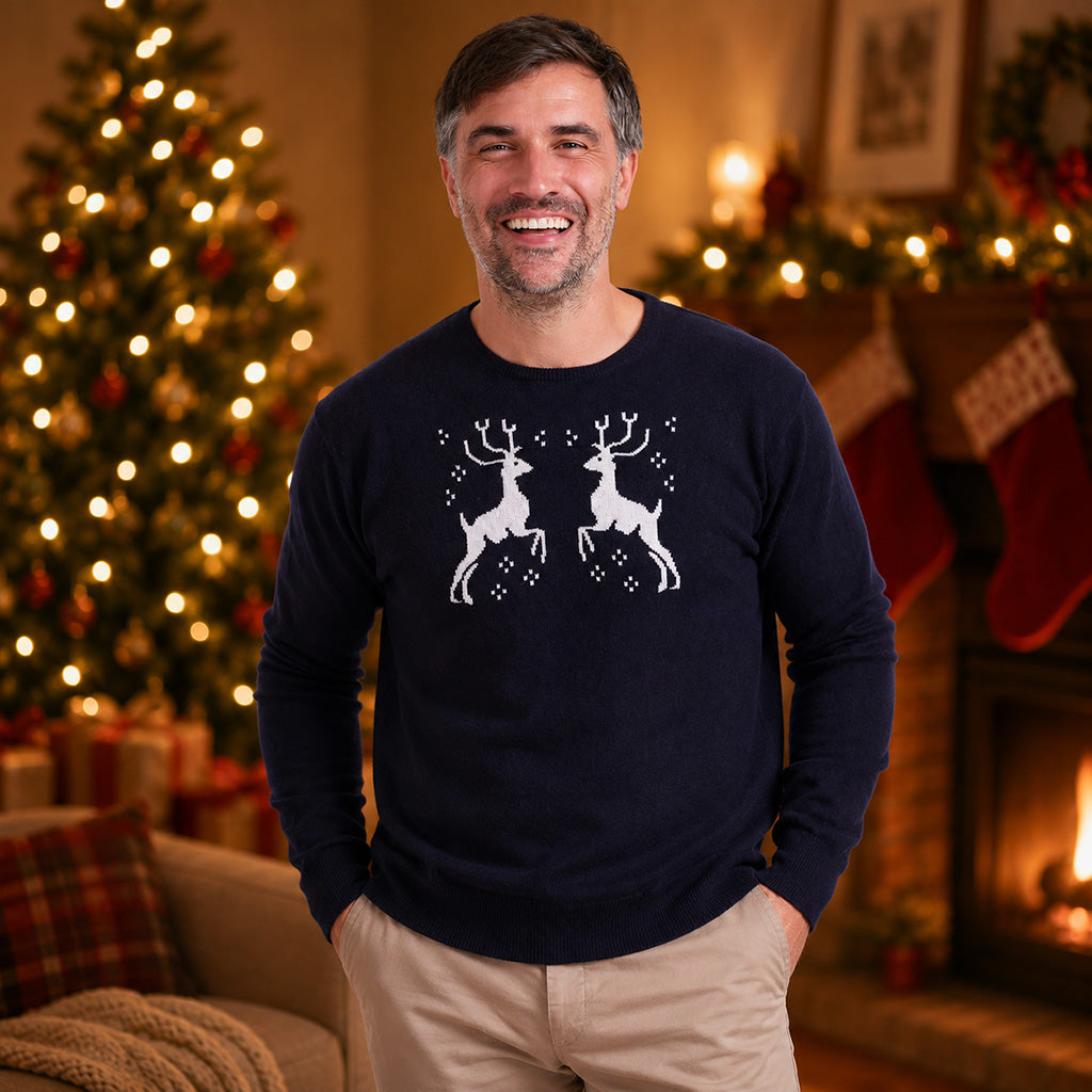 A man wearing a cashmere navy blue crewneck sweater with a white graphic of two leaping reindeer, standing in a cozy, warmly lit living room with a Christmas tree and fireplace in the background.