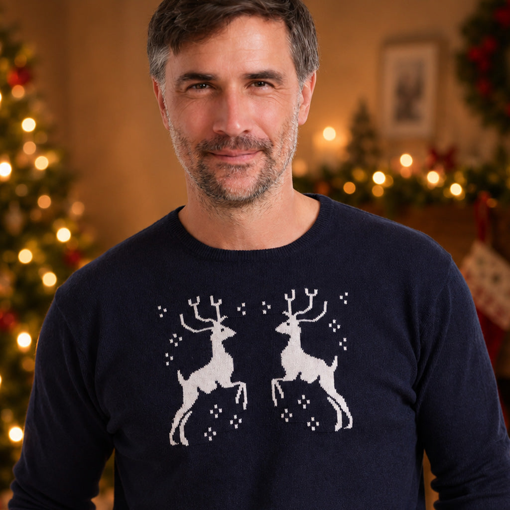 close up of a man wearing a cashmere navy blue crewneck sweater with a white graphic of two leaping reindeer, standing in a cozy, warmly lit living room with a Christmas tree and fireplace in the background.