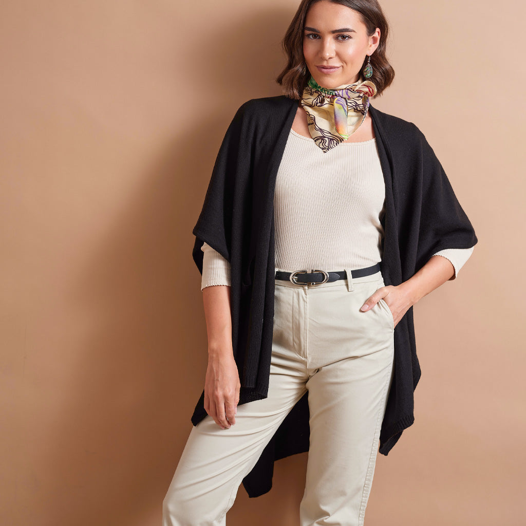 A woman models a black open-front cape cardigan over a cream ribbed knit top and cream pants, accessorized with a patterned neck scarf and a black belt, set against a solid tan studio background.
