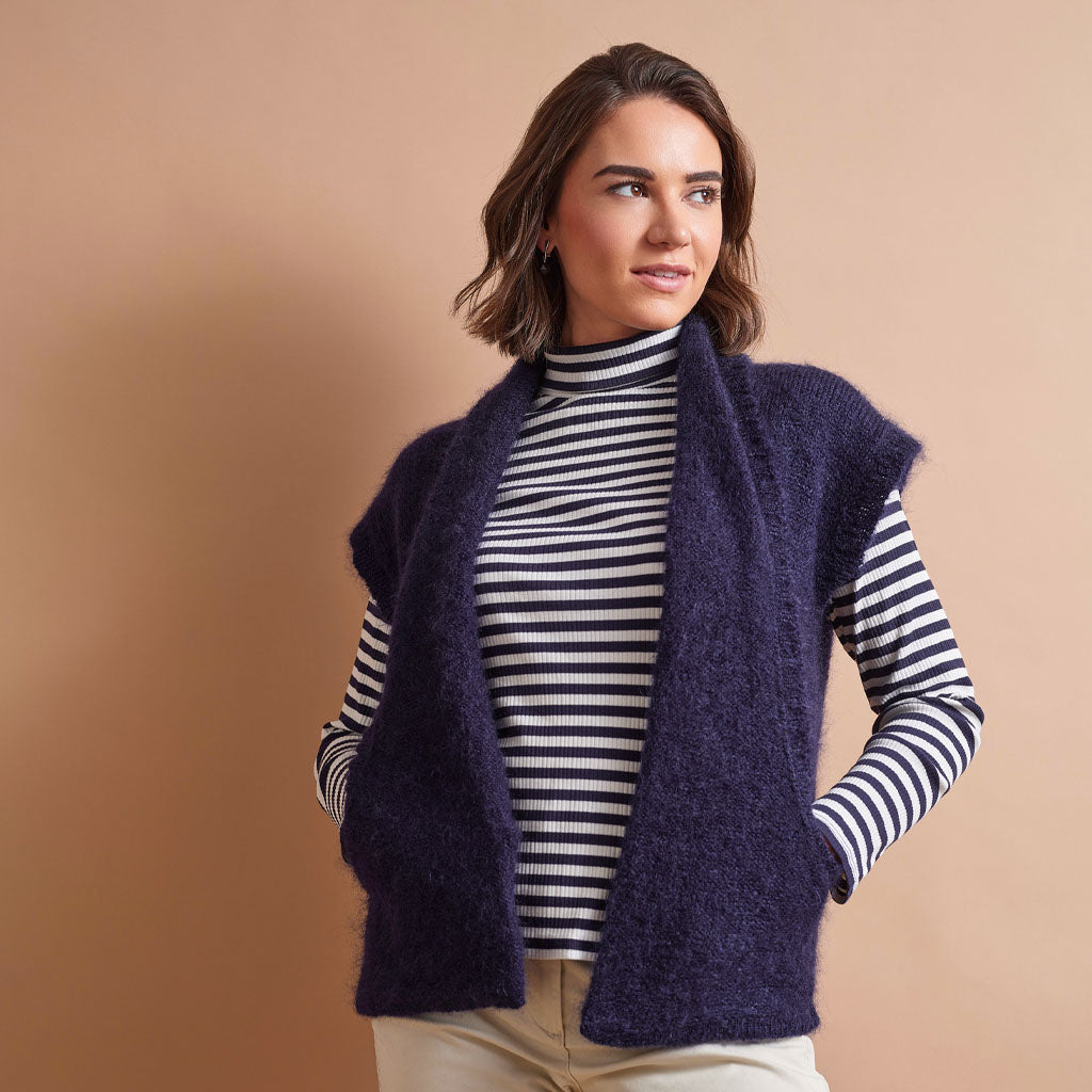 A woman wearing a fuzzy dark purple open-front vest over a navy and white striped turtleneck sweater, paired with light beige pants, set against a solid warm beige background.