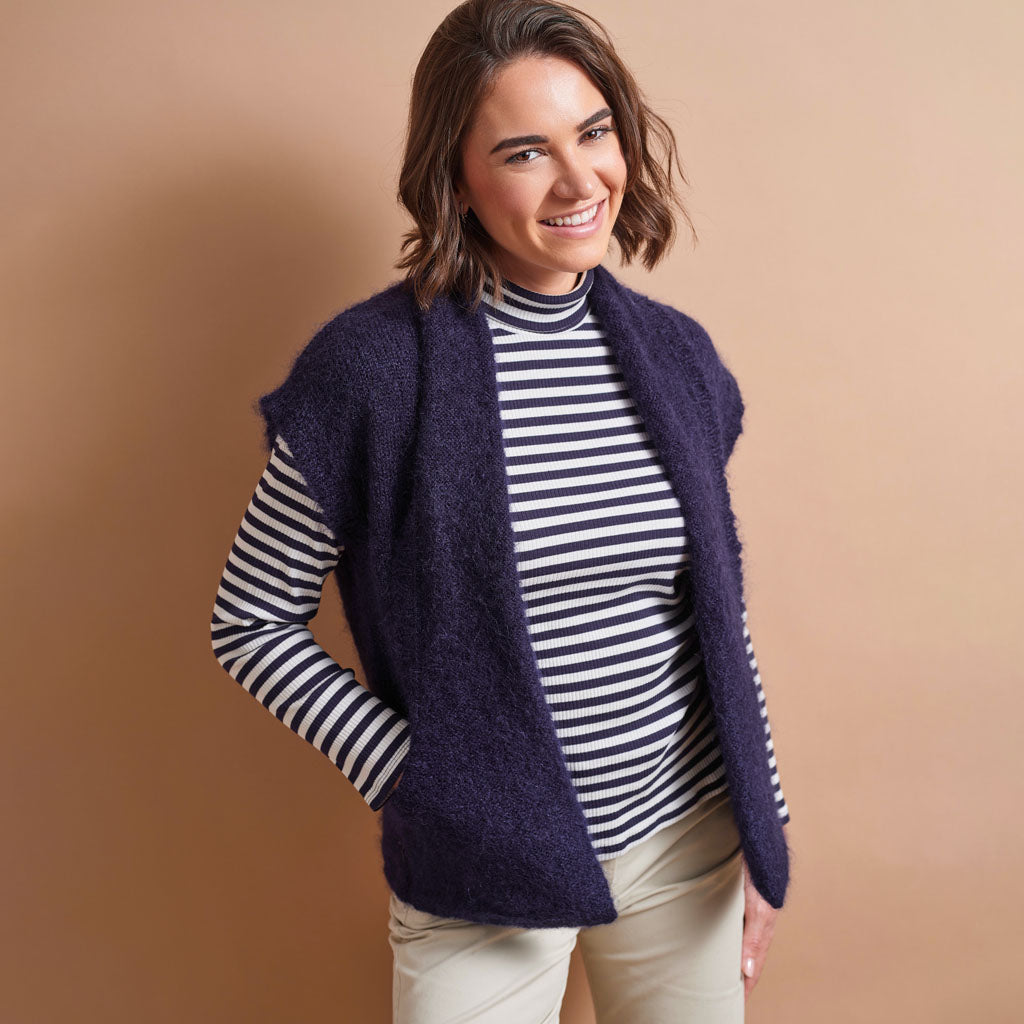A woman modeling a fuzzy, sleeveless dark navy knit vest over a black and white striped turtleneck, paired with beige pants, against a solid warm tan studio background.